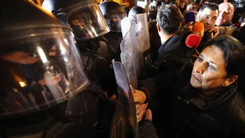 Getty Images Salome Zourabichvili menghadapi barisan polisi anti huru hara bertopeng. Polisi anti huru hara mengenakan balaclava di bawah helm anti huru hara dan memegang perisai anti huru hara. Kerumunan mengelilingi presiden. 