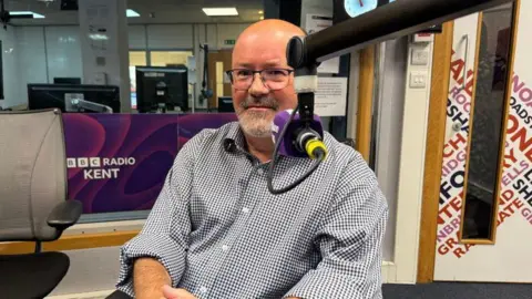 A man sat in a radio studio with a radio mic partially covering his face. He is wearing a blue and white shirt and a pair of glasses. 