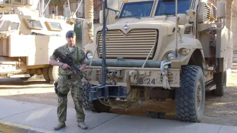 Steve Hooper Steve is wearing combats, boots and an RAF beret. He is holding a rifle and standing in front of a military truck