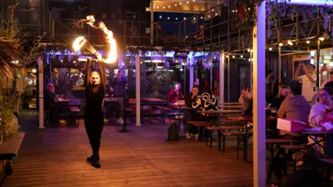 SPARK:York A fire artist performs, twirling burning sticks in the air. Around them, people sit at tables among shipping containers.