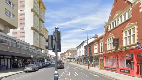 Street View image showing a road flanked by shops. Tower blocks rise over the shops to the left, there is a traffic light and a pedestrian crossing in the centre of the image, at the forefront.