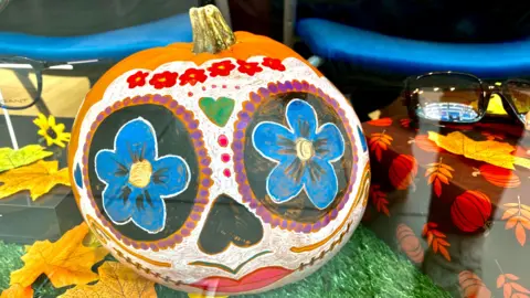 A pumpkin that has had a face painted on it, with blue flowers for eyes, an upside down heart for a nose and pink lips. The face of the pumpkin has been painted white and there is a row of red flowers along the top.