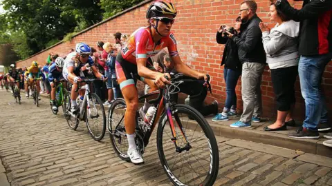 Getty Images Cyclists racing up a cobbled street, with people standing on a path cheering them on.