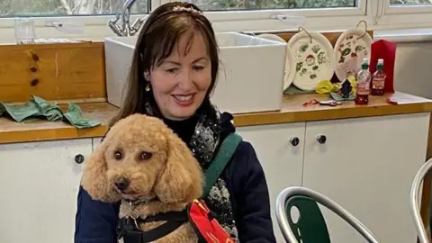 A woman sat with a dog that is wearing a harness. She has long dark hair and is smiling down at the dog.