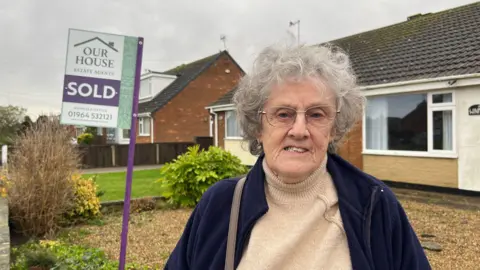 Amanda White/ BBC A head and shoulder shot of Janet Hall smiling at the camera stood in front of a bungalow with a "SOLD" house sign in the background. She has short curly grey hair and is wearing glasses, a beige roll-neck sweater and a blue zip-up fleece. 