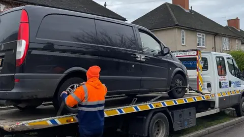 A man wearing a hi-vis jacket and an orange hat with his back to the camera is strapping the rear wheels of a black van to the back of a car transporter. The car transporter has a white cab. There are houses in the background.