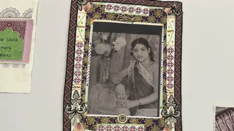 Julita Waleskiewicz A framed black‑and‑white portrait is displayed within an ornate handmade textile border, alongside a small sign reading “One World, many cultures".