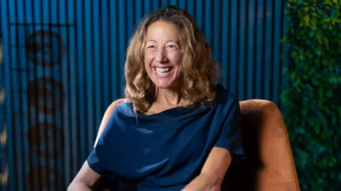 Supplied A woman sat on a brown chair, smiling just off camera. She is wearing a blue dress. She is sat in a blue room.