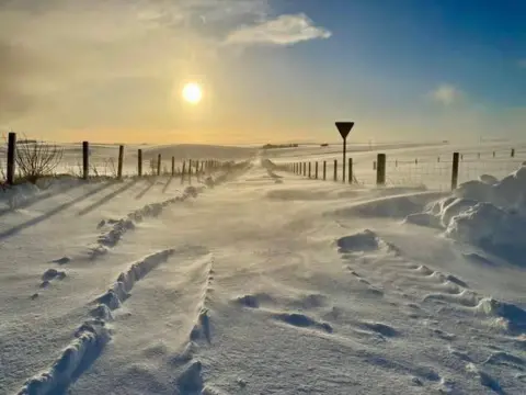 A rural road covered in snow with fences either side. The sun is low in the sky.