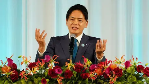 Takeharu Yamanaka in a suit addresses an audience from a decorated podium filled with vibrant flowers against a curtain backdrop