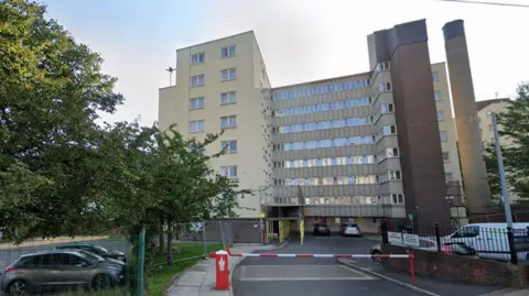 View of a tower block, consisting of eight storeys. The is a barrier on the entrance road and cars parked to either side.