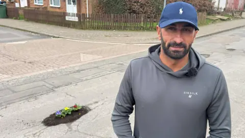 BBC A man in a grey hoodie and cap standing infont of a pothole he's filled in with flowers and soil. 
