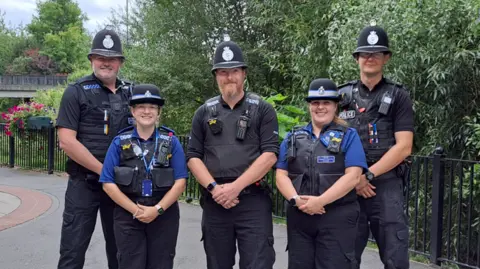 West Mercia Police A group of five police officers, three men and two women, stand together in a pedestrian area and smile for the camera.