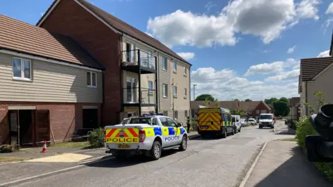 A police car and police forensics van parked outside some houses on Davies Road in Salisbury. Police tape surrounds one of the properties.  