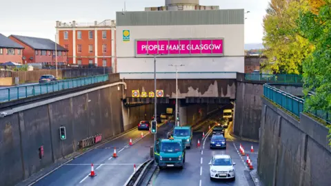 Traffic entering and leaving the Clyde Tunnel in Glasgow. A large People Make Glasgow sign hangs above it. 