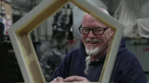 A man with glasses and a long white beard in a ponytail laughs as he tinkers with a wooden model in a workshop 