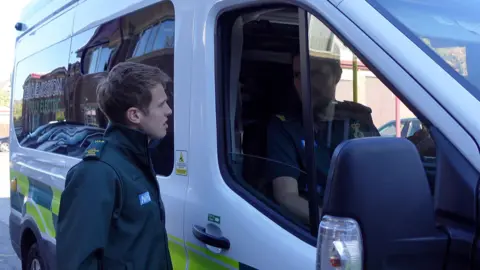 Corbishley next to one of the ambulances at Yorkshire Ambulance Service HQ in Wakefield. He's wearing a dark green uniform and speaking to someone inside the ambulance