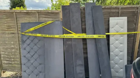Gabby Colenso/BBC Parts of a bed, including a grey velour headboard, stand against a wooden fence. Around them is yellow crime scene tape.