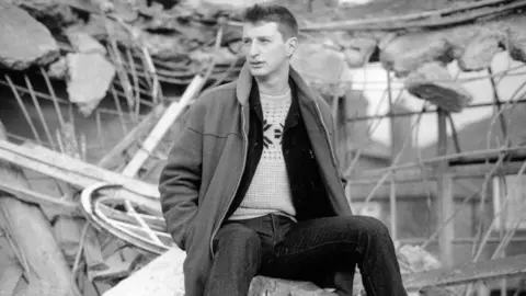 Getty Images Black and white image shows a young Billy Bragg sitting in the rubble of a building, looking to the left of the frame. He has his hands in the pockets of a long coat. 