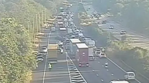 The view from a traffic camera. Traffic is standstill and queued all the way along a motorway. There are three lanes and a slip road on the left hand side where the queues are, and on the right hand side there are also three lanes with traffic flowing. 