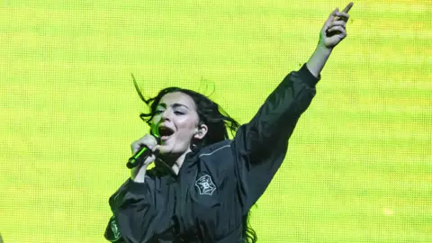 Charli XCX performing in London. Charli is a woman in her early 30s with long curly black hair worn loose. She wears a black boiler suit and sings into a microphone, her left hand pointing skywards. She's in front of a neon green backdrop
