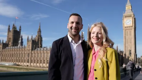 PA Media Hannah Spencer, wearing a mustard-yellow coat, poses for photos at outside the House of Commons with Green Party leader Zack Polanski