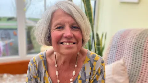 Linda Joyce-Jones is a white woman in her 60s, she was white hair in a bob cut and is sitting on her sofa with a multi-coloured knitted blanket over the top of it. She is wearing a grey and yellow dress and a necklace with white beads on. Her garden is visible in the background over her shoulder