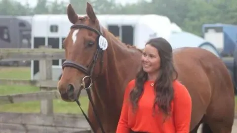 Family Photo A woman standing next to a brown horse. She is wearing a peach jumper with a gold necklace around her neck. The woman has brown hair and has her hair in a half up half down style. She is smiling at the horse. They are standing outside next to a brown fence. 
