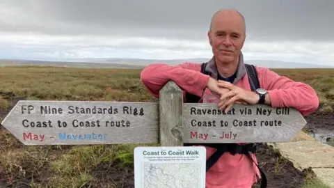 Dr Chris Elliot is wearing a pink jumper and is perched on a wooden sign giving directions in the countryside. 