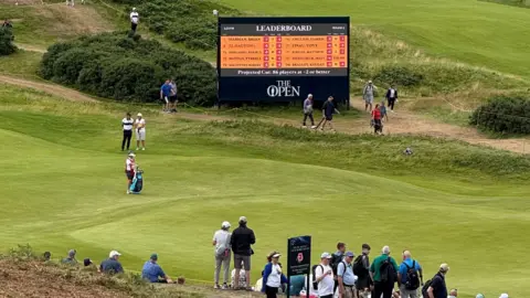 Royal PortraSh é uma tela grande com tabela de classificação aberta em campo aberto. Existem várias pessoas em pé no campo de golfe. 