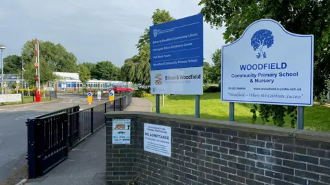 The entrance to a primary school car park, as indicated by a sign saying 'Woodfield Community Primary School & Nursery'. 