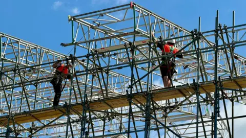 Cornerstone Kettering Two men, in high-vis outfits, with helmets on, standing on a platform, taking down scaffolding. There is a wooden platform. A blue sky is behind them. 