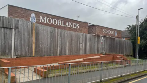 Tony Fisher/BBC The entrance to "Moorlands School" which has a path and steps leading up to it. The school is behind a wooden fence. In the foreground there is a footpath and railings.