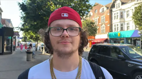 Richard Knights/BBC Max Barrett is wearing a red beanie hat, glasses, a white T-shirt and a gold chain in Clacton.