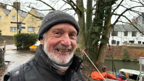 Martin Heath/BBC Alan Scarisbrick with grey hair and beard, smiling at the camera while wearing a thick black coat and grey woollen hat. He is standing by a river, with a tree immediately behind him. A burnt-out boat is visible to the right of the picture. There is a dark-coloured signpost to the left of the picture in front of a yellow building.
