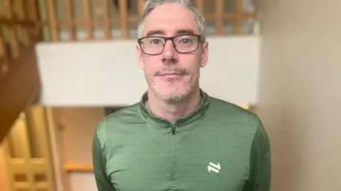 Thomas is standing in a room. He wears glasses and has a green top on with a white logo. The background is blurred.