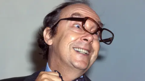 Eric Morecambe clowns for the camera wearing his glasses off centre.
