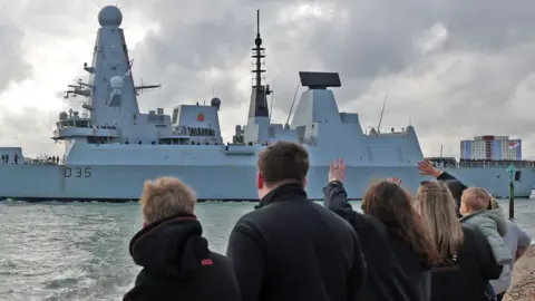 People display a banner and wave at the Royal Navy destroyer HMS Dragon on the day it departs, heading to the Eastern Mediterranean to bolster British defences in the region, in Portsmouth, Britain, March 10, 2026.