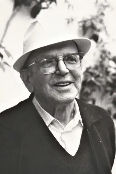 Letizia Chiocchetti A black and white photograph of an elderly man in glasses and a white hat. He is wearing a shirt and a V-neck jumper. He is smiling and looking to the left of the camera. 