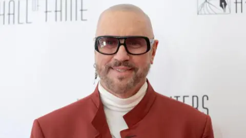 Getty Images A bald man with grey facial hair wearing one earring and sunglasses, as well as a white polo kneck jumper and red jacket.