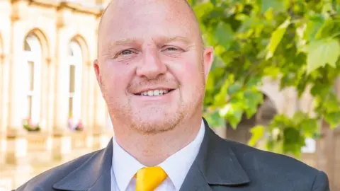 LDRS A bald man with light facial hair wearing a grey suit jacket, white shirt and yellow tie.
