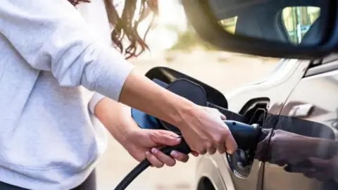 Getty Images A woman fills up an electric car. Her hands are visible on the pump. She is wearing a grey jumper. The car is black.