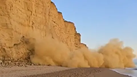 Suzanne Sears Cliffs to the left of a beach with a large cloud of rubble tumbling onto the beach.