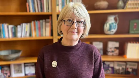 Karen MacKinnon has short, bobbed, blonde hair and is wearing glasses. She is wearing a burgundy high-neck jumper with a gold circular brooch. 