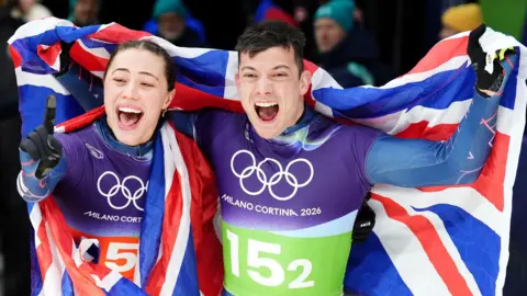 PA Media Tammy Stoecker and Matt Weston celebrate after winning gold. Matt Weston is holding up a Union Jack flag behind him and raising their arms in the air.