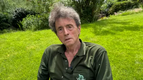 A man with greying wavy hair sits outdoors on a garden chair. He is wearing a dark green polo shirt with a small embroidered crest on the chest. Green lawn, shrubs and mature trees fill the background.