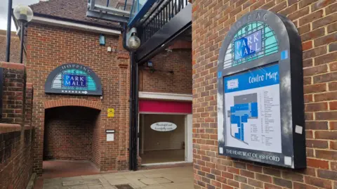 Liz Saul/BBC An entrance to an outdoor shopping centre. There is a large map on one wall showing the plan of the centre and in the back ground, an empty shop, with an old sign in the glass window.