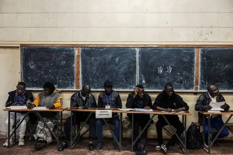 AFP via Getty Images A group of officials sit behind trestle tables. Behind them are four school blackboards fixed to the wall.