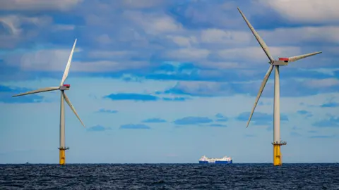 Two offshore wind turbines with white towers and yellow bases stand in the sea - a ship can be seen in the background, passing between them 
