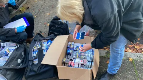 Boxes of illegal cigarettes and tobacco in a box and two black holdalls on the pavement